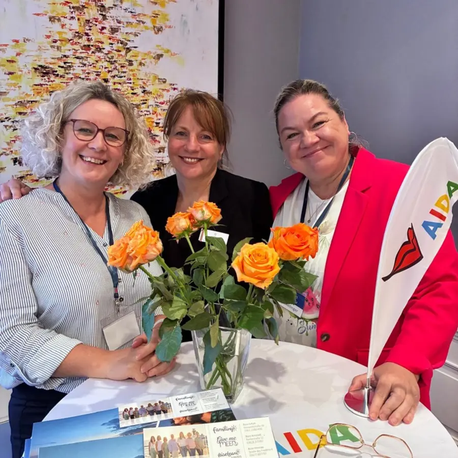 Drei lächelnde Kolleginnen mit Rosen am Tisch mit AIDA Flagge